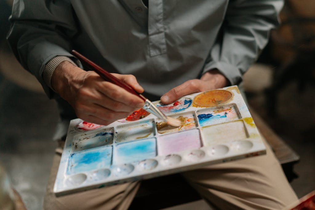 Close-up of an artist's hands mixing colors on a watercolor art palette with a paintbrush.
